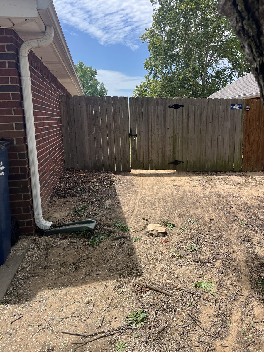 A downspout tied into buried drainage pipe with the trench backfilled along a brick wall to the fence line.
