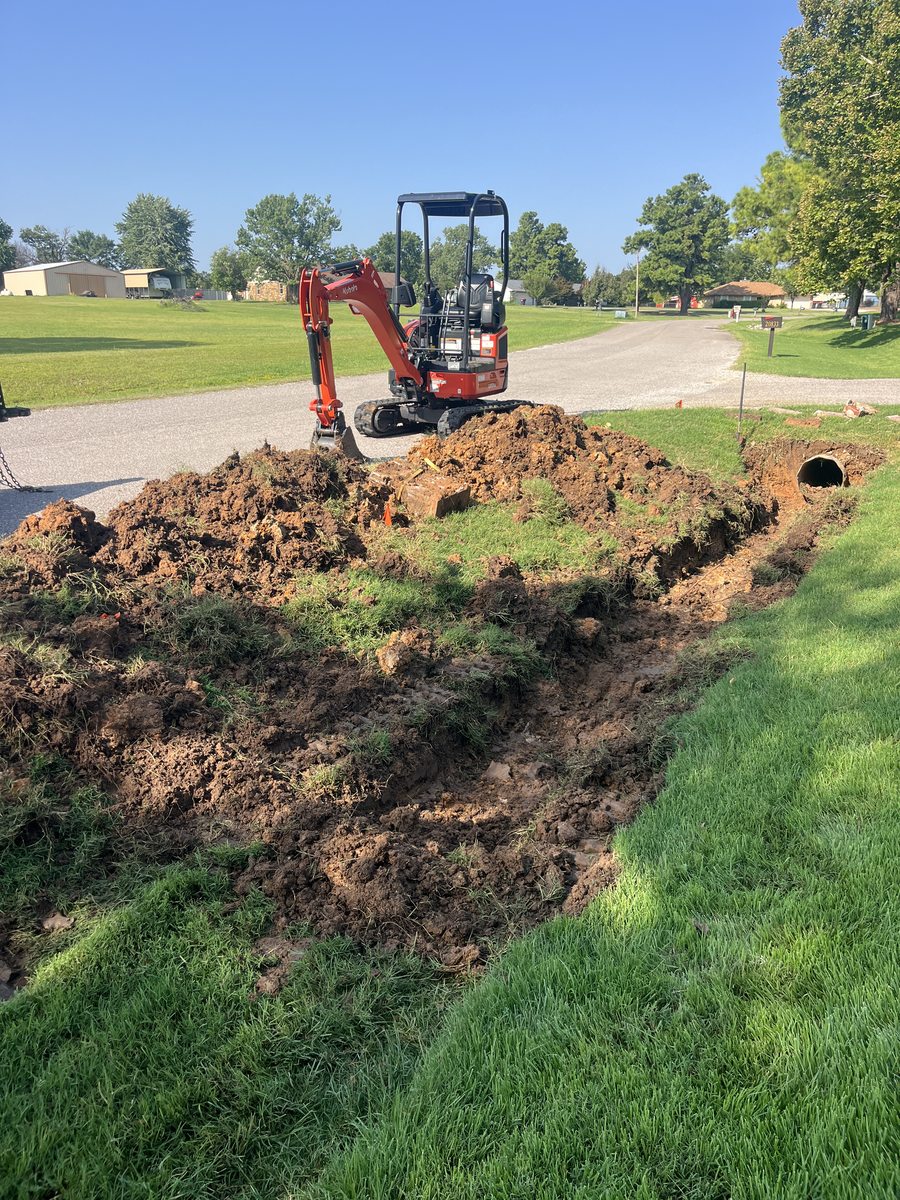 A Kubota mini excavator with an open trench and a corrugated culvert pipe ready to set along a rural driveway.