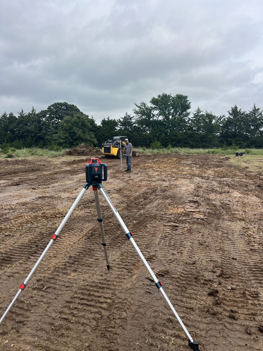 A rotary laser level on a tripod with a crew member holding a grade rod beside a skid steer on a freshly cut pad.